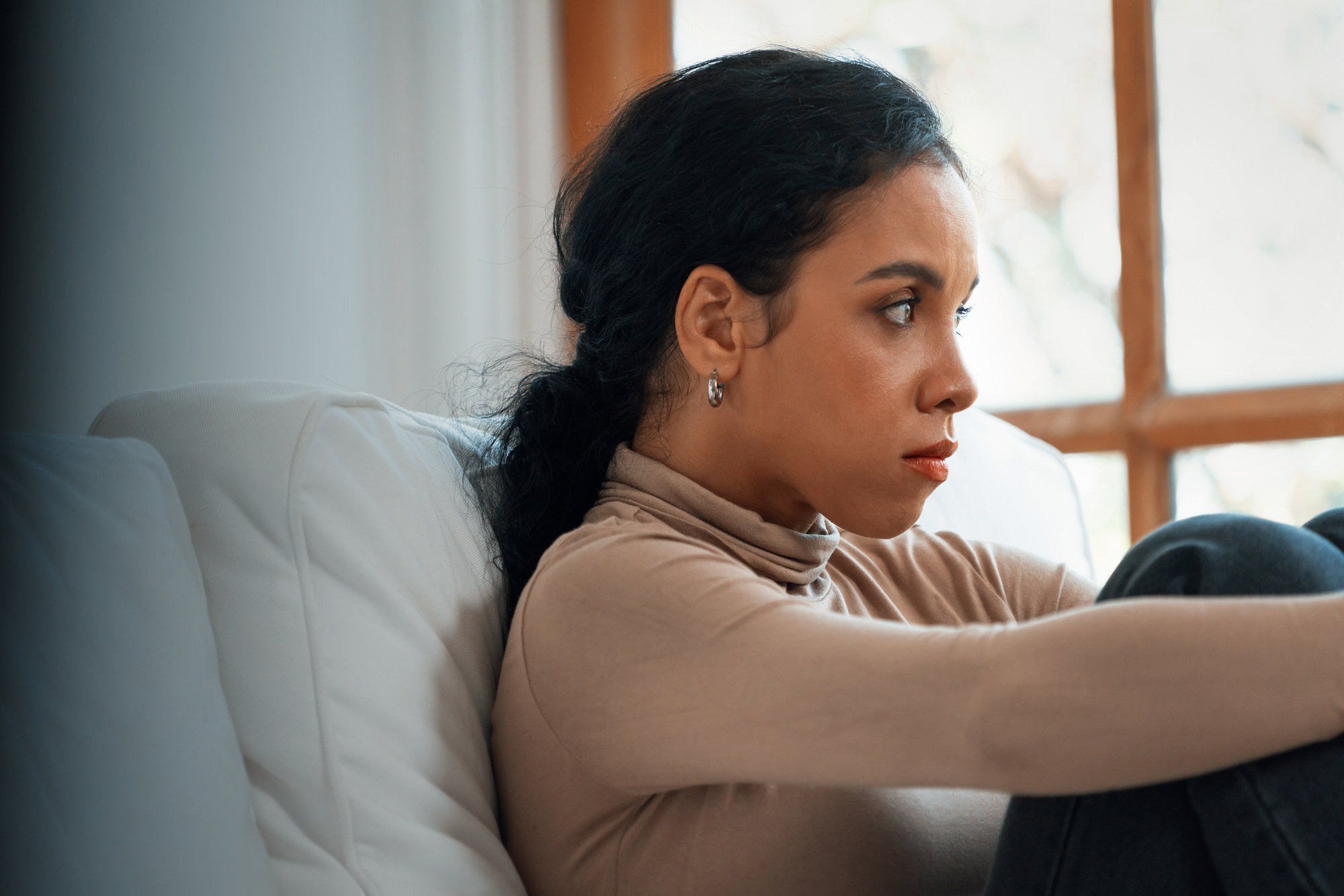 A woman with long dark hair sits on a white sofa, gazing thoughtfully out a window. She is wearing a beige turtleneck and appears relaxed yet deep in thought in a softly lit room.