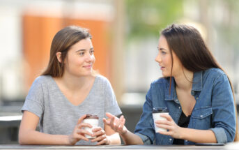 Two women are sitting at an outdoor table, each holding a takeaway coffee cup. They are engaged in conversation, with one gesturing as she speaks. The background is softly blurred, suggesting a casual, relaxed setting.