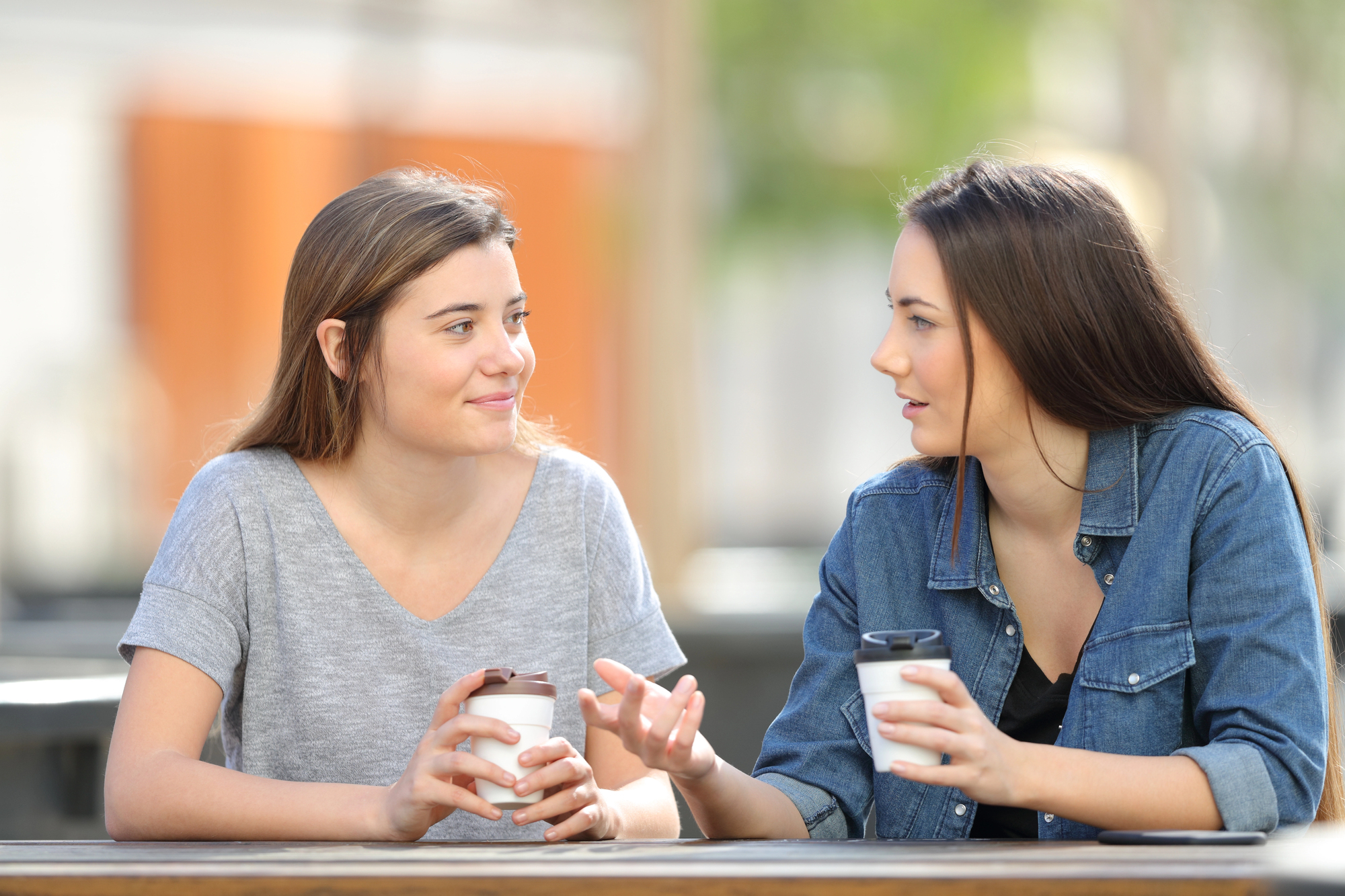 Two women are sitting at an outdoor table, each holding a takeaway coffee cup. They are engaged in conversation, with one gesturing as she speaks. The background is softly blurred, suggesting a casual, relaxed setting.