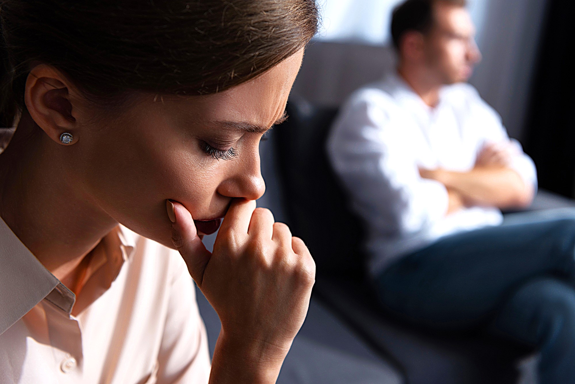 A woman appears deep in thought, resting her chin on her hand, while a man sits in the background with his arms crossed, looking away. Both seem to be in a contemplative or tense moment on a sofa.