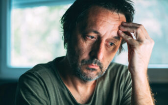 A man with short, messy hair and a beard is sitting indoors, looking pensive. He rests his head on his hand, with a window blurred in the background. He wears a green shirt and appears deep in thought.