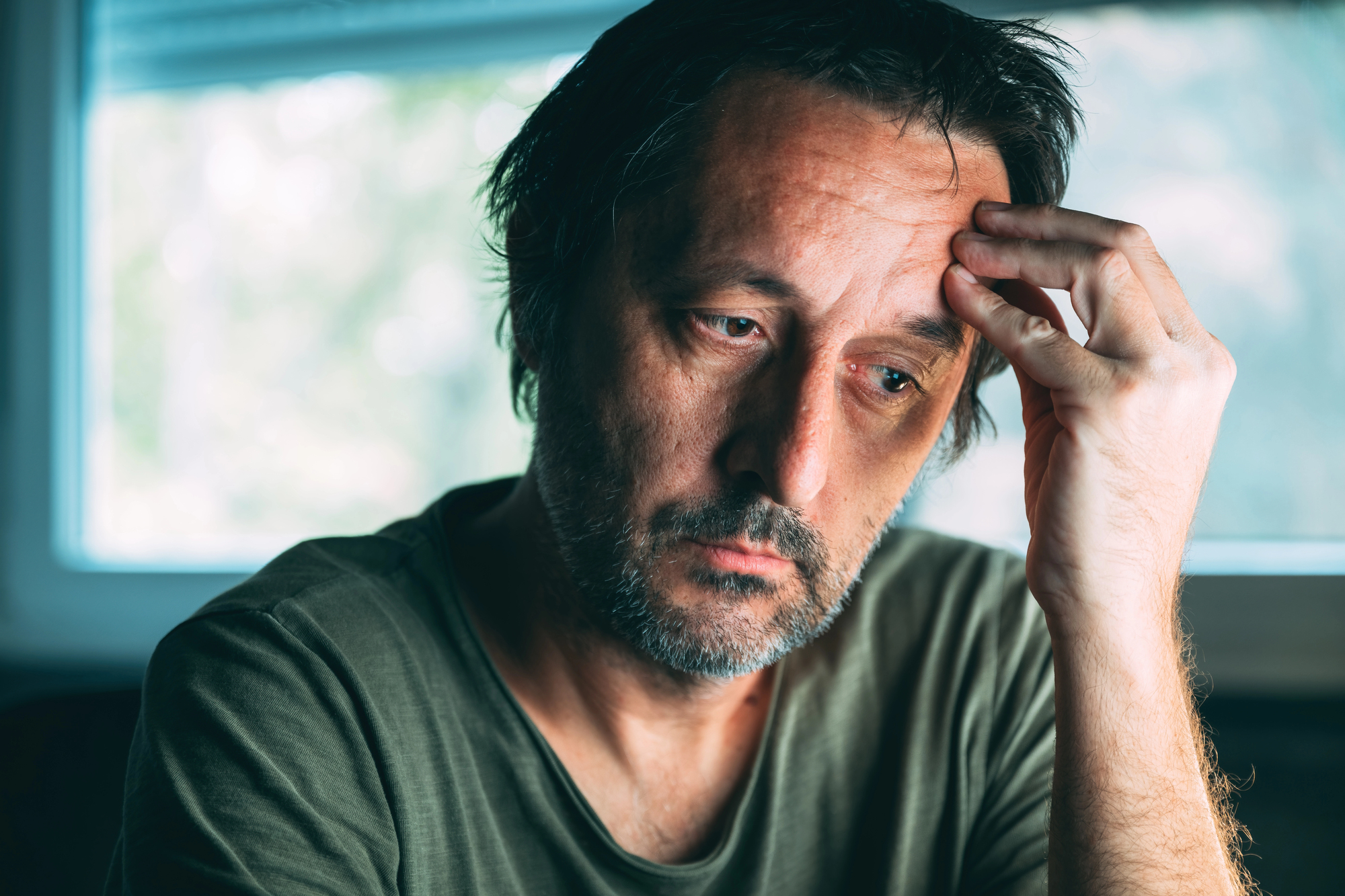 A man with short, messy hair and a beard is sitting indoors, looking pensive. He rests his head on his hand, with a window blurred in the background. He wears a green shirt and appears deep in thought.