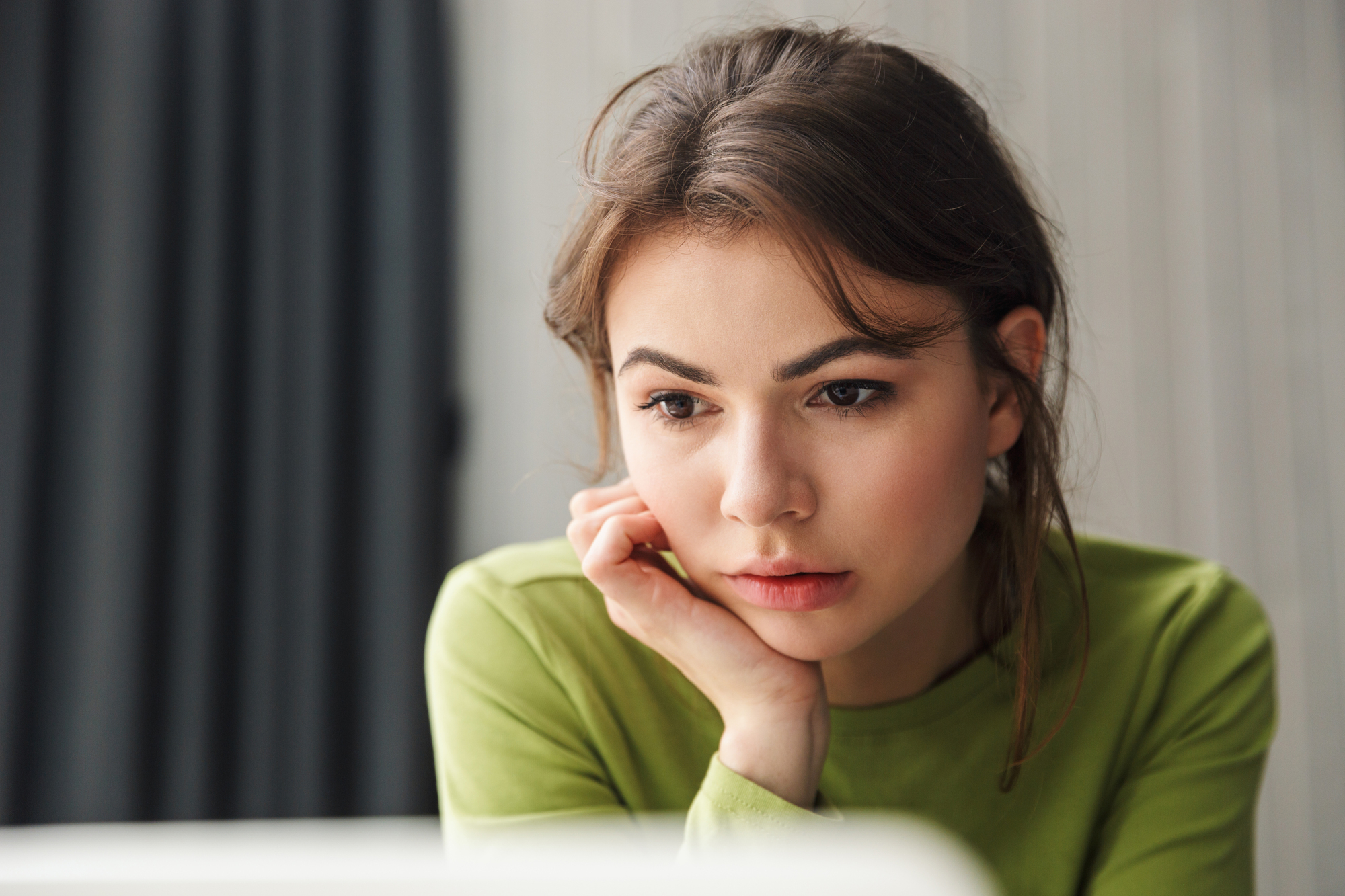 A woman with long brown hair wearing a green shirt rests her chin on her hand, gazing intently at something off-screen. The background is softly blurred, suggesting an indoor setting.