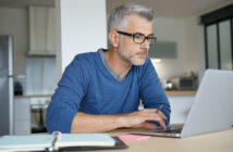 A man with glasses and gray hair works intently on a laptop at a table in a modern kitchen. He is wearing a blue sweater, and there is an open notebook with a pen nearby.