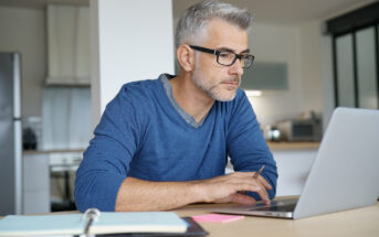 A man with glasses and gray hair works intently on a laptop at a table in a modern kitchen. He is wearing a blue sweater, and there is an open notebook with a pen nearby.