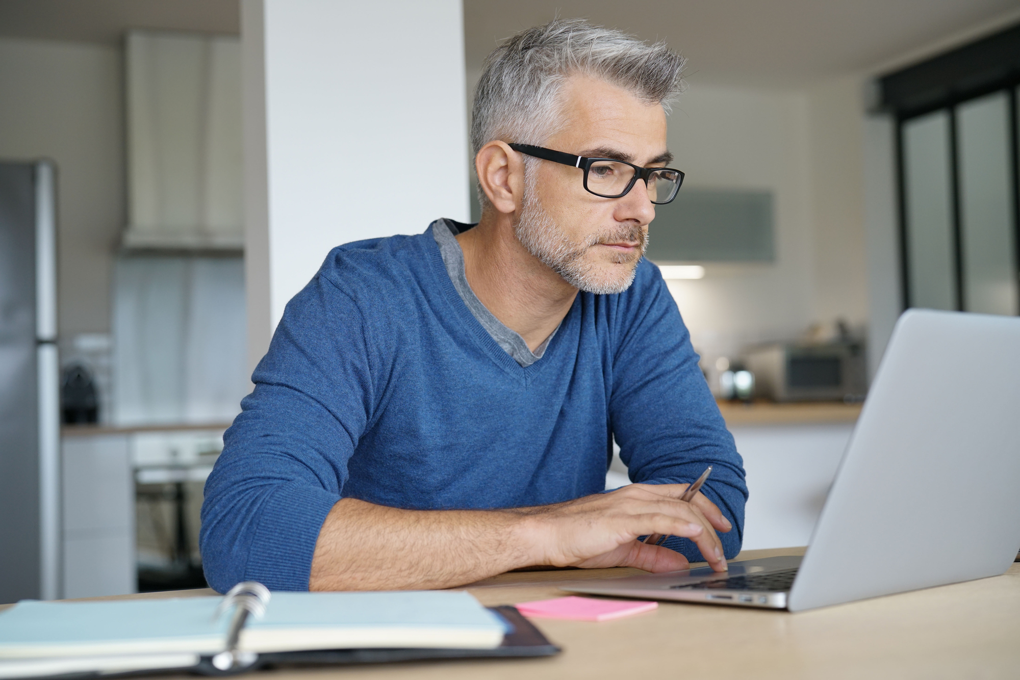 A man with glasses and gray hair works intently on a laptop at a table in a modern kitchen. He is wearing a blue sweater, and there is an open notebook with a pen nearby.