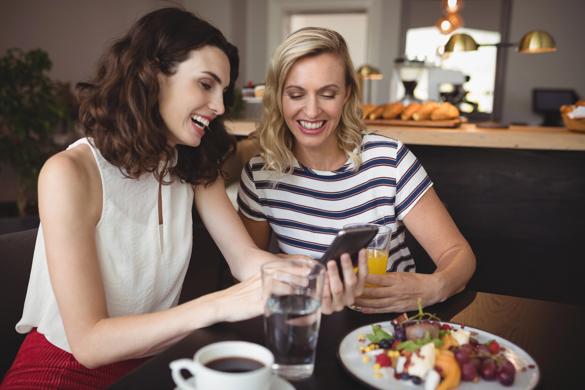 Two women sitting at a cafe table, smiling and looking at a smartphone. They have drinks and a plate of food with fruit and salad in front of them. The background shows a counter with pastries under warm lighting.