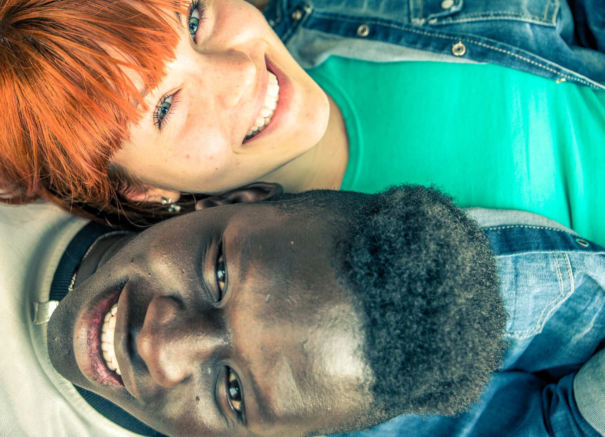 Two people lying head to head on the ground, smiling at the camera. The person with red hair is wearing a green shirt and blue denim jacket, while the person with short curly hair is dressed in a white shirt and also has a denim jacket.