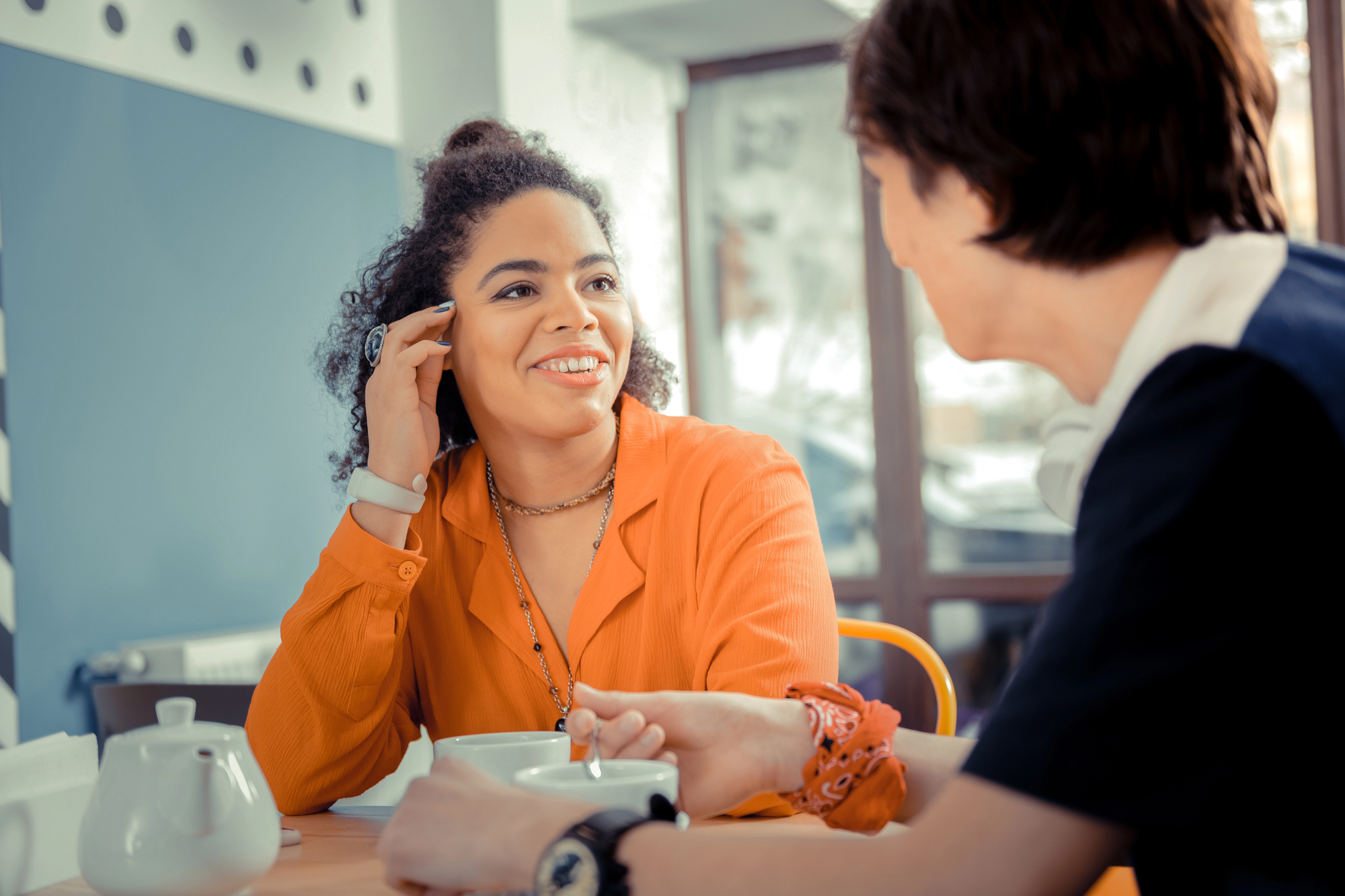 Two people are sitting across from each other at a table in a bright, airy café. One person is wearing an orange shirt and smiling, while the other person, facing away from the camera, is engaged in conversation. A teapot and cups are on the table.