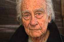 An elderly man with white hair and a weathered face is looking directly at the camera. He is wearing a dark jacket, and the background is a wooden texture. His expression is contemplative and serene.