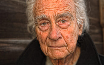 An elderly man with white hair and a weathered face is looking directly at the camera. He is wearing a dark jacket, and the background is a wooden texture. His expression is contemplative and serene.