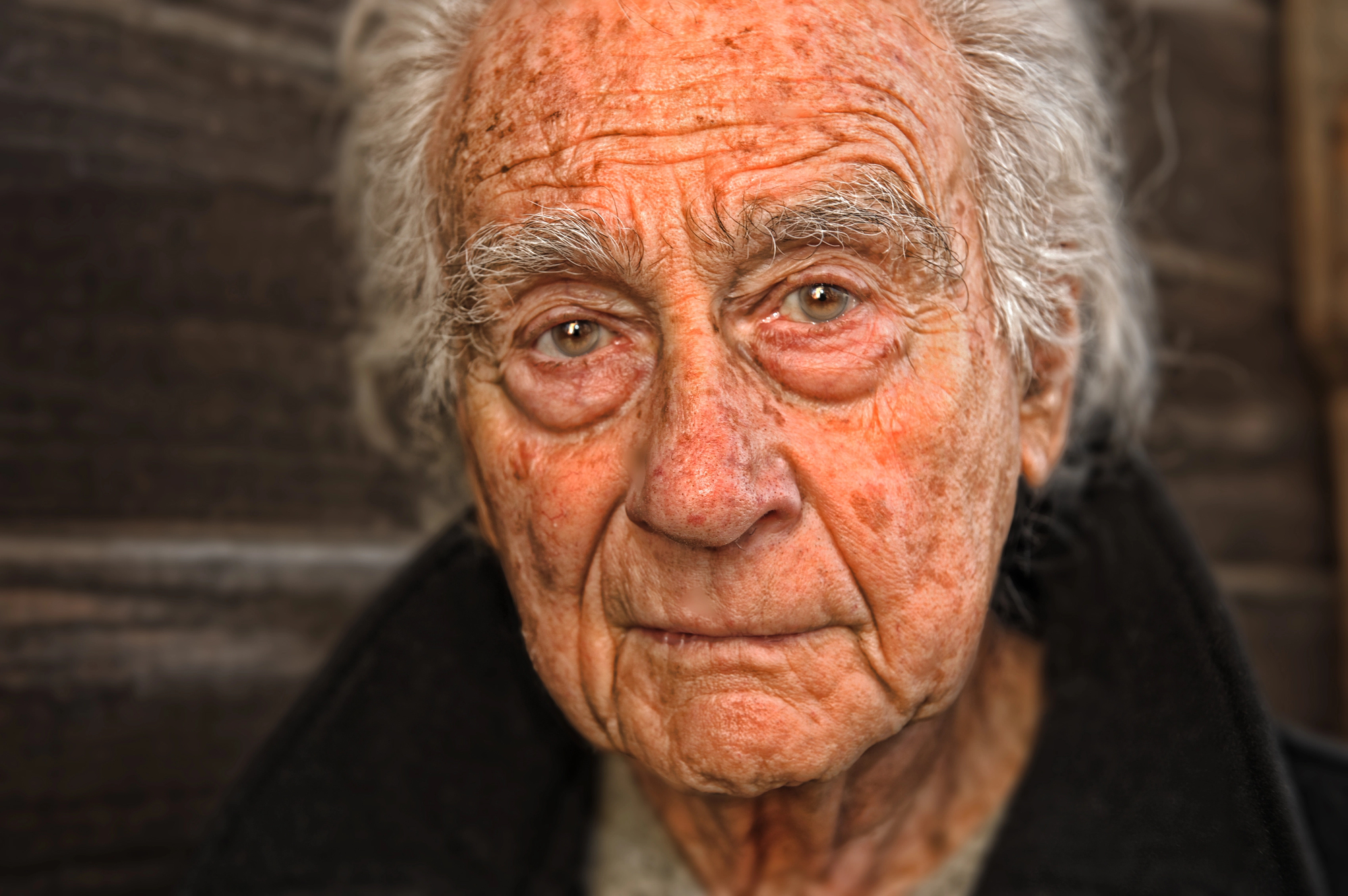 An elderly man with white hair and a weathered face is looking directly at the camera. He is wearing a dark jacket, and the background is a wooden texture. His expression is contemplative and serene.