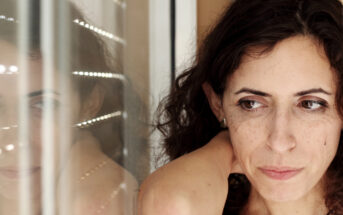 A woman with curly hair looks pensively to the side. She is standing indoors near a window with blinds partially open, casting a soft light across her face. Her reflection is faintly visible in the glass.