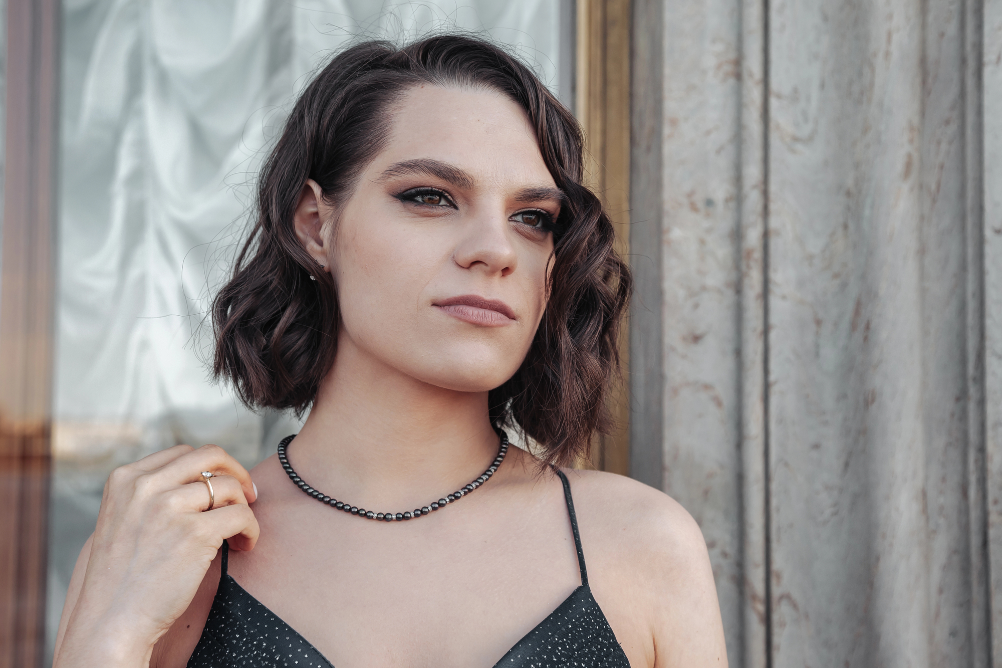 A person with shoulder-length dark hair stands in front of a stone wall, wearing a black, sleeveless top and a beaded necklace. They look off into the distance with a thoughtful expression.