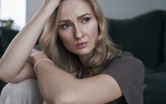 A woman with long blonde hair sits on a sofa, looking contemplative. She rests her head on one hand, with the other arm wrapped around her knees, wearing a short-sleeve brown top in a softly lit room.