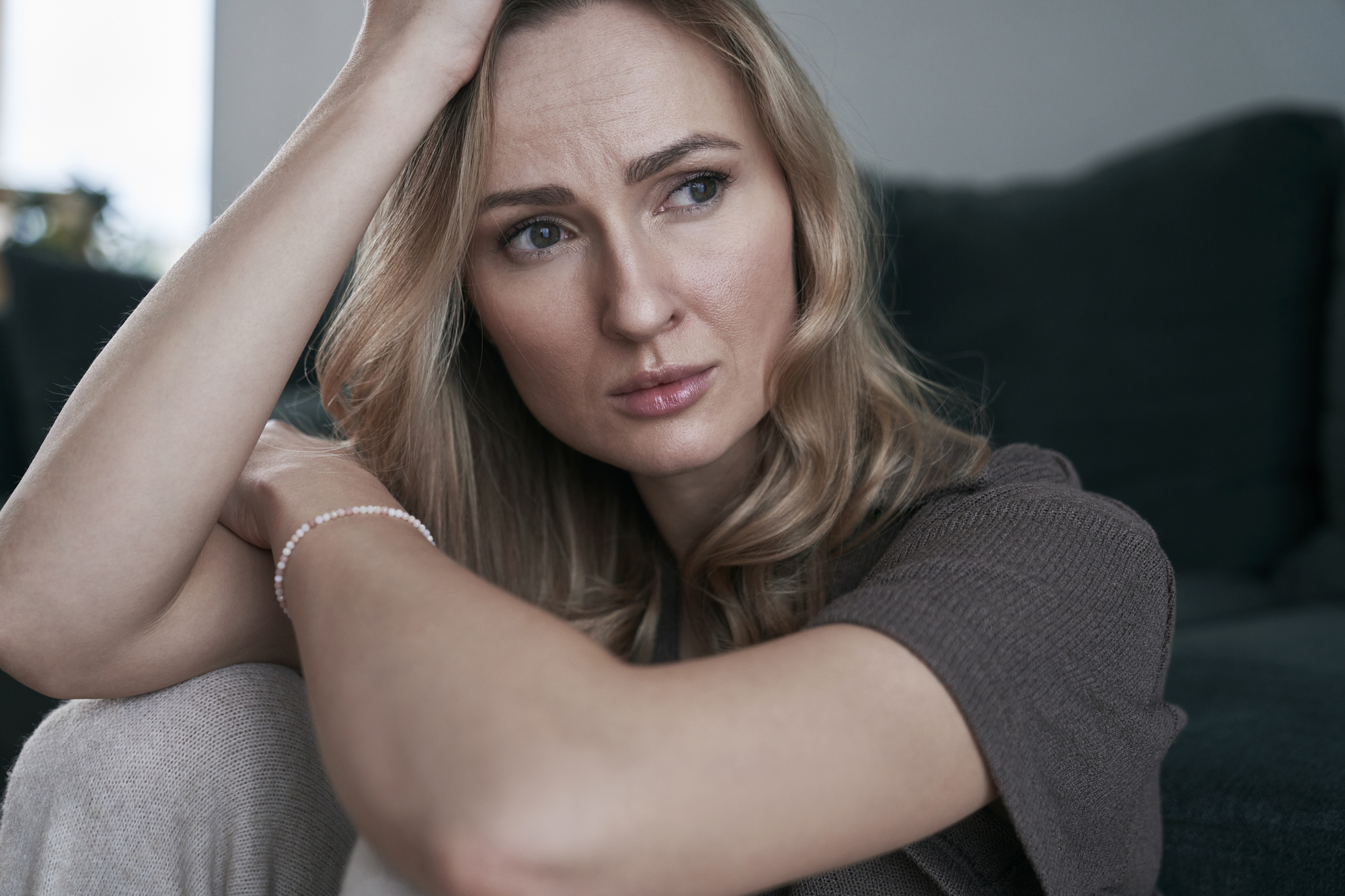 A woman with long blonde hair sits on a sofa, looking contemplative. She rests her head on one hand, with the other arm wrapped around her knees, wearing a short-sleeve brown top in a softly lit room.