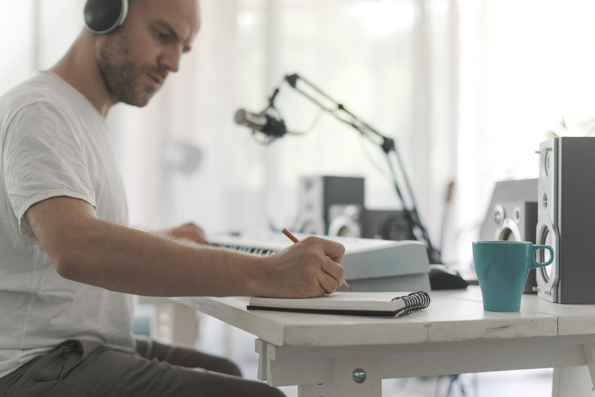 A person wearing headphones sits at a desk with a keyboard, writing in a notebook. A microphone, speakers, and a blue coffee mug are on the table. The background is softly lit, suggesting a home studio environment.