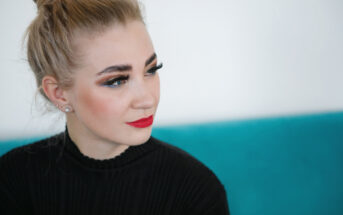 A woman with a topknot hairstyle and bold makeup, including eyeliner and red lipstick, gazes to the side. She wears a black top and sits against a turquoise background.