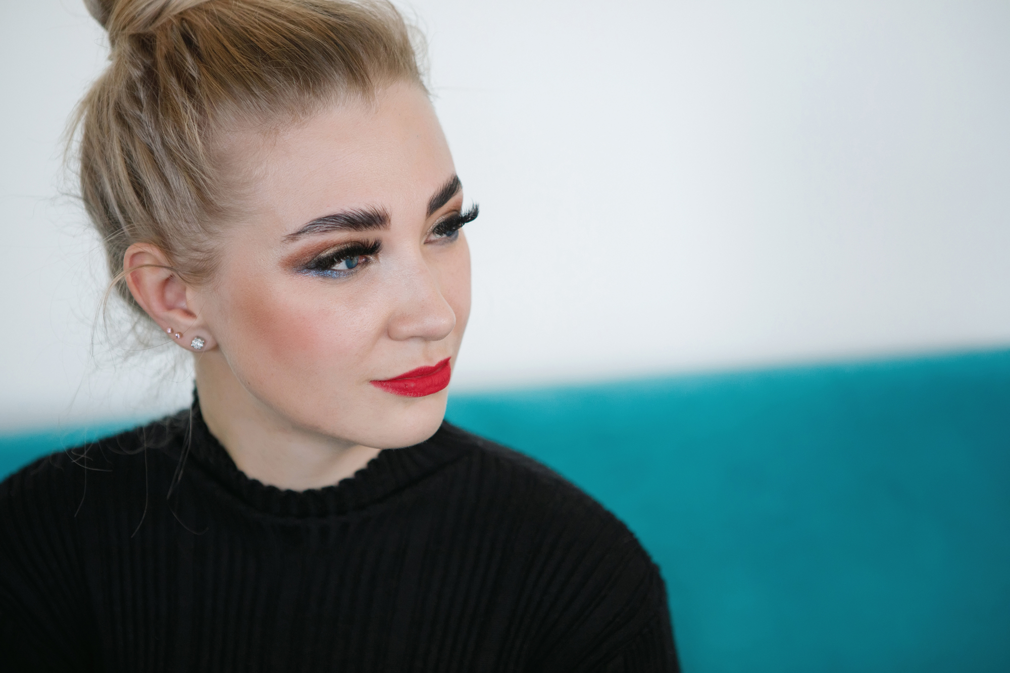 A woman with a topknot hairstyle and bold makeup, including eyeliner and red lipstick, gazes to the side. She wears a black top and sits against a turquoise background.