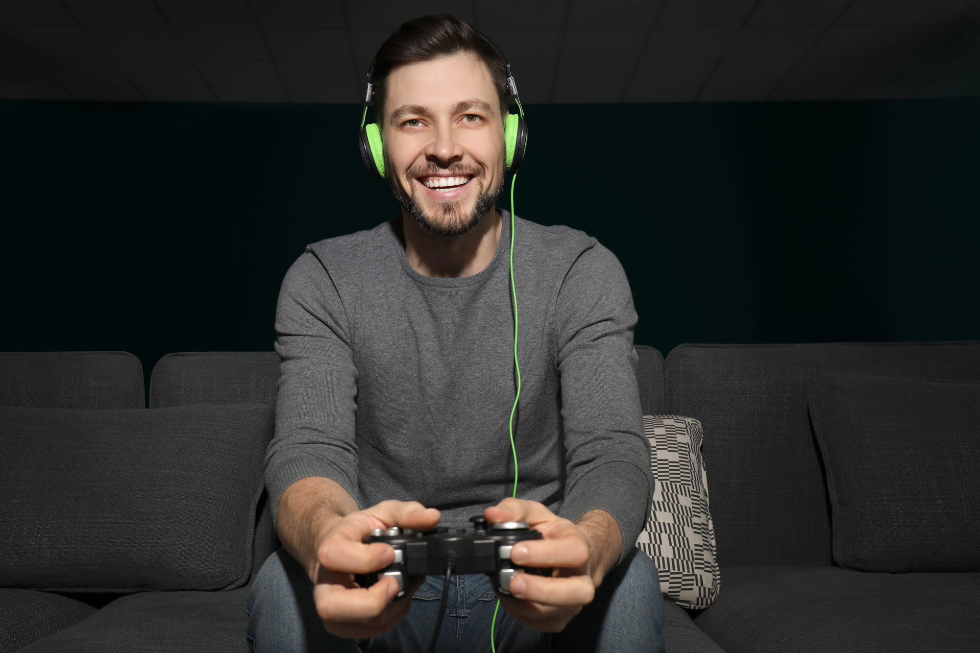 A man with a beard is sitting on a couch, holding a video game controller. He is wearing a gray sweater and green headphones, smiling while engaged in playing a game. The background is dark, enhancing the focus on him.