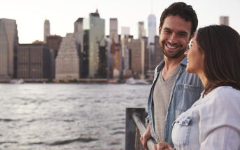 A man and a woman smile at each other while standing on a waterfront railing. Tall city buildings are visible in the background across the water, under a clear sky.