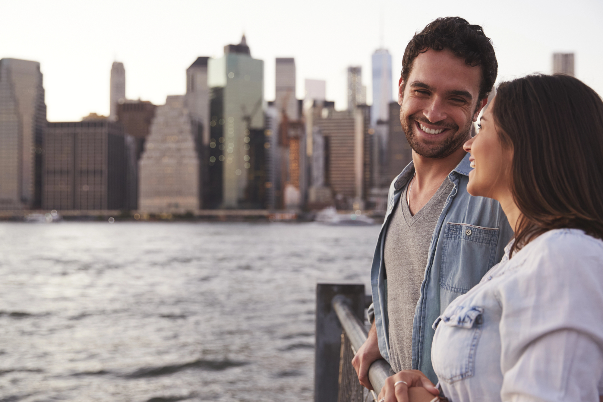 A man and a woman smile at each other while standing on a waterfront railing. Tall city buildings are visible in the background across the water, under a clear sky.