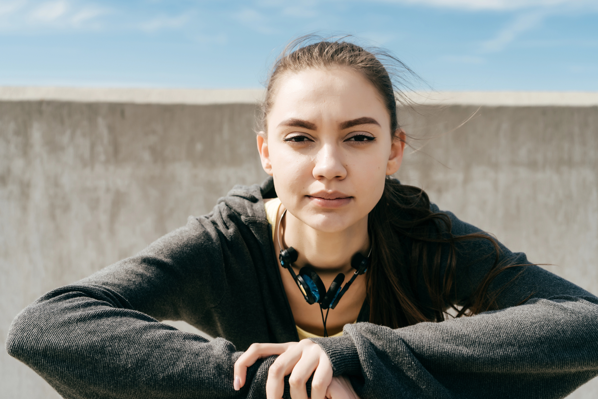 A young woman with long hair and wearing a dark hoodie sits outdoors. She has earphones around her neck and is looking directly at the camera with a neutral expression. A blue sky and a concrete wall are in the background.