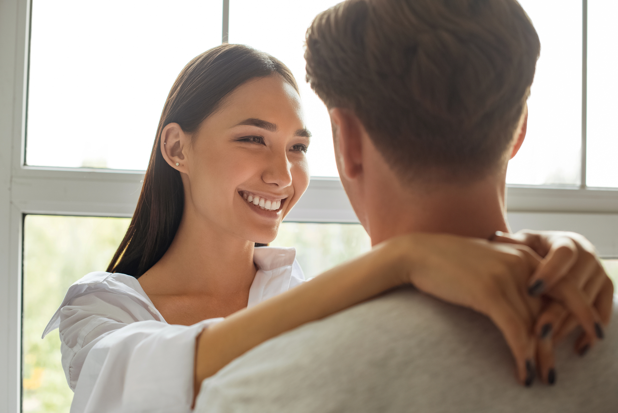 A woman smiling and hugging a man indoors by a window. The woman has long dark hair and is wearing a white shirt, while the man has short hair and is wearing a light-colored shirt.