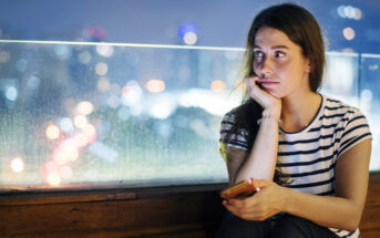 A woman in a striped shirt sits on a bench, holding a phone and looking thoughtful. The background shows a blurry cityscape with colorful lights at night, visible through a transparent barrier.