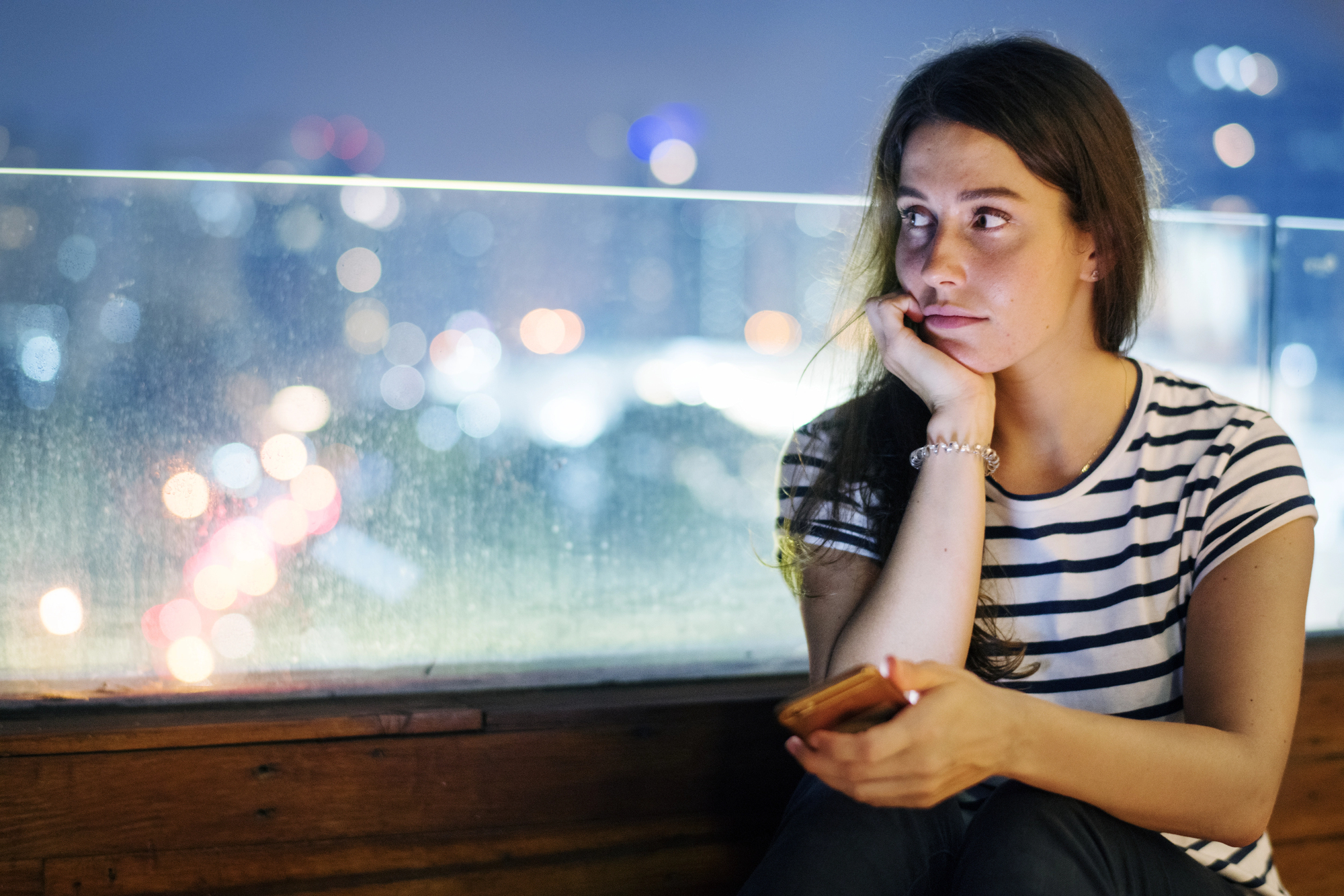 A woman in a striped shirt sits on a bench, holding a phone and looking thoughtful. The background shows a blurry cityscape with colorful lights at night, visible through a transparent barrier.