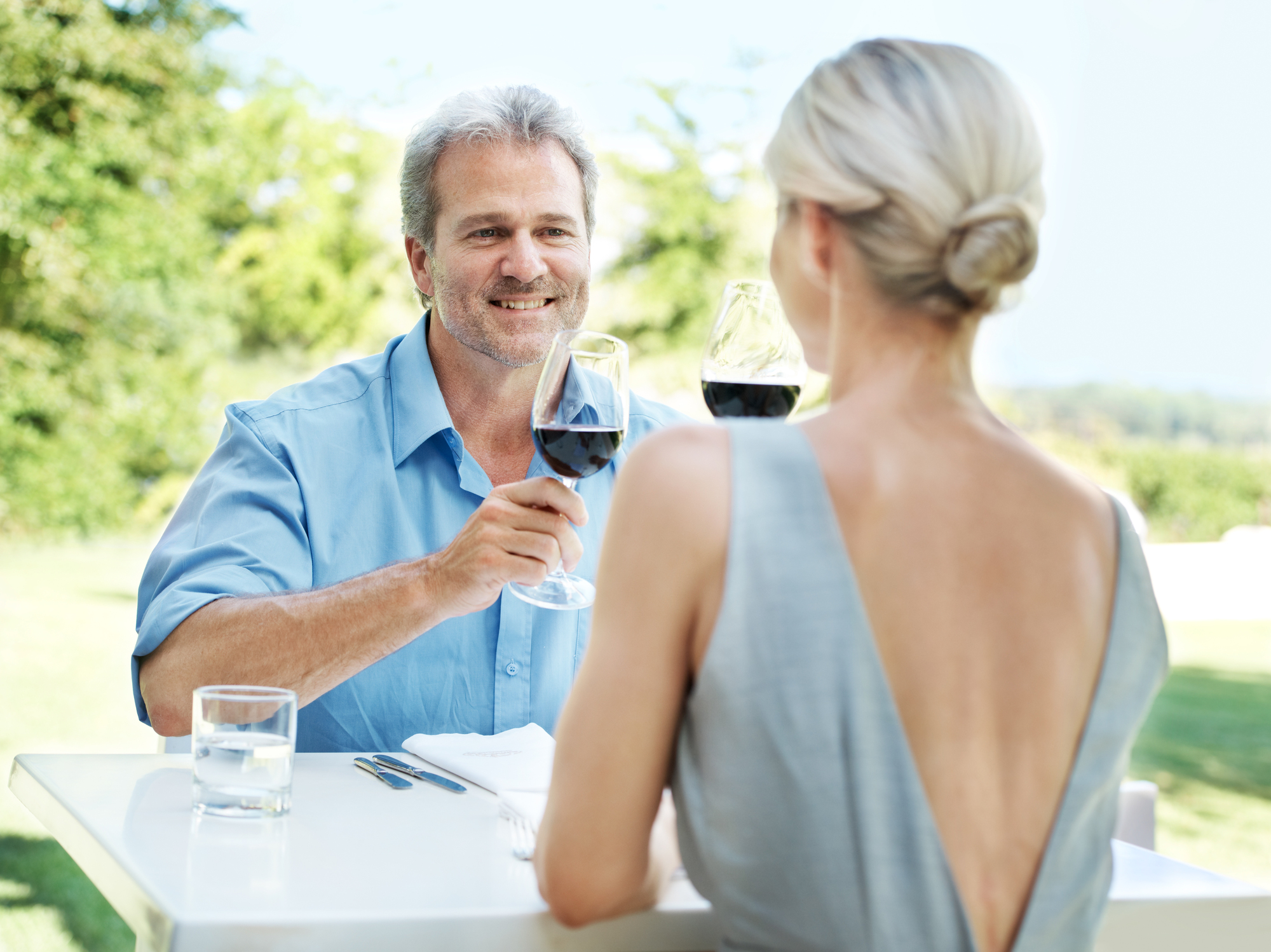 A man and woman sit at an outdoor table, toasting with glasses of red wine. The man wears a blue shirt, and the woman is in a backless dress. They are smiling, with greenery in the background.