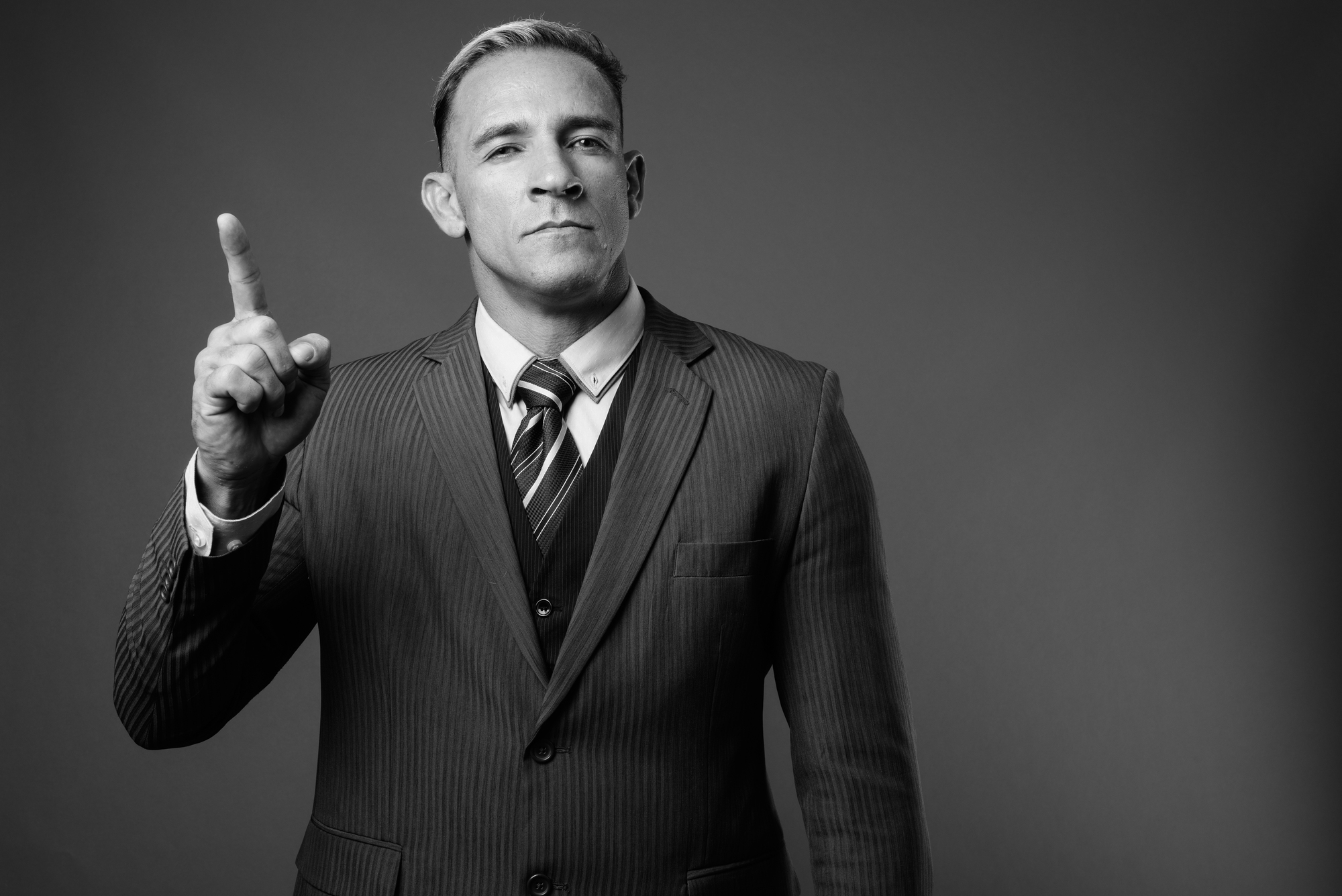 A man in a suit, with a serious expression, raises his index finger. The image is in black and white, giving it a formal and classic look. The background is plain, emphasizing the man's gesture and attire.