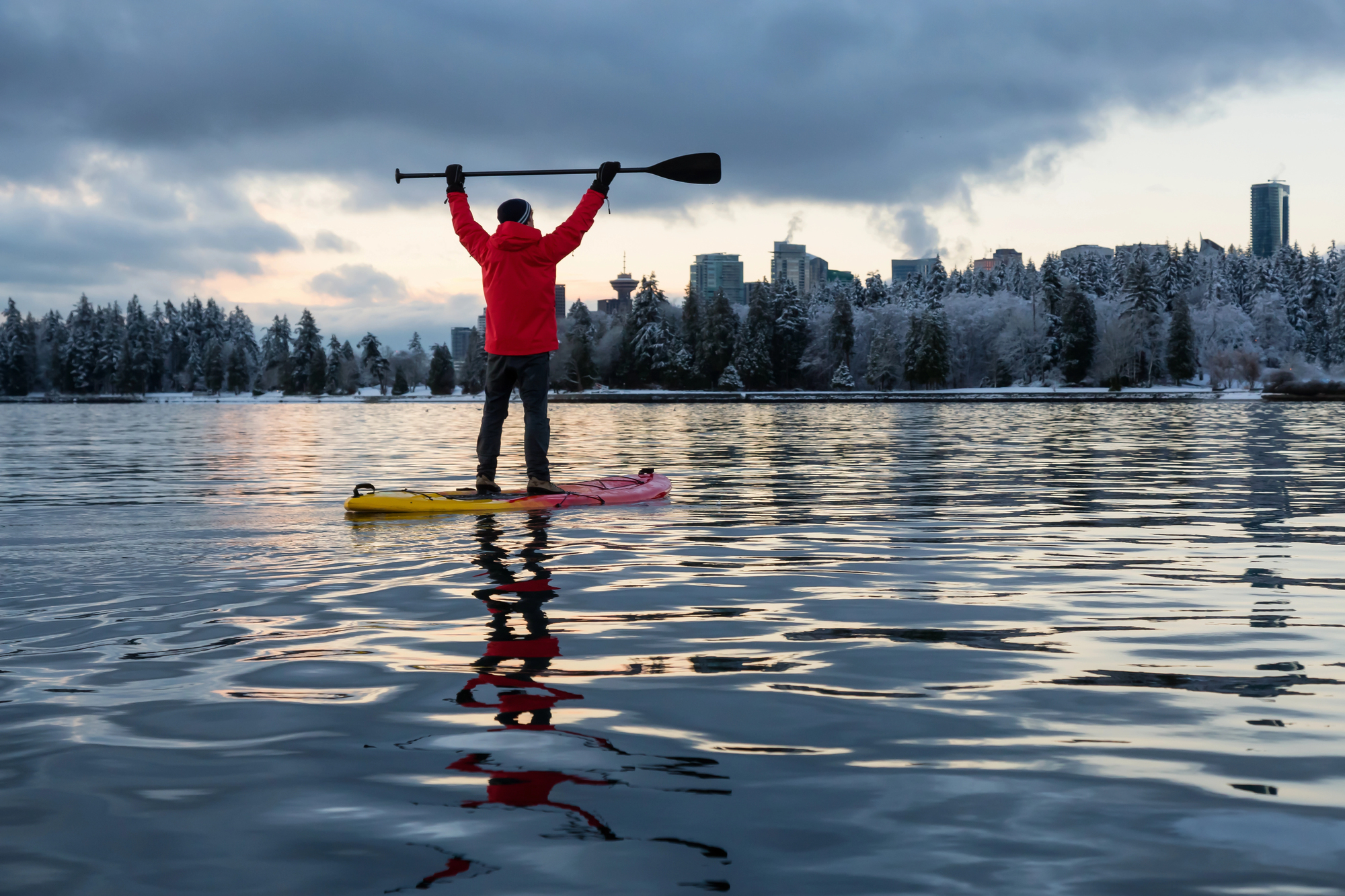 A person in a red jacket stands triumphantly on a paddleboard, holding a paddle overhead. They are on a calm body of water with a backdrop of snow-dusted trees and city buildings under a cloudy sky.