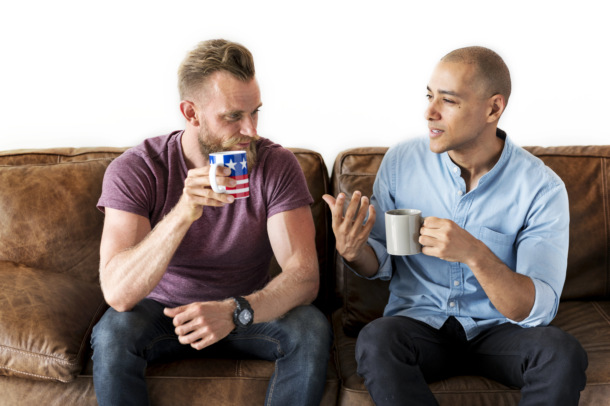 Two men sit on a leather couch engaged in conversation, each holding a mug. One wears a maroon shirt and holds a mug with a flag design, while the other wears a light blue shirt with a plain mug, gesturing with his free hand.