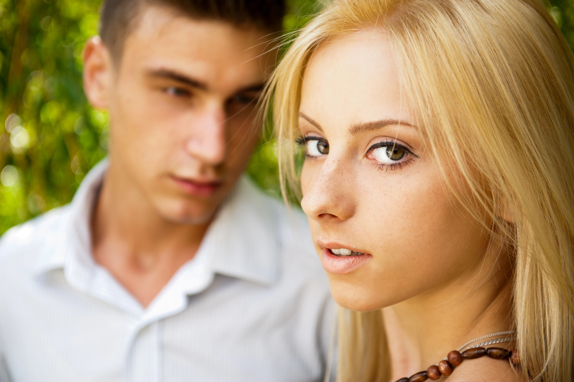 A close-up of a young blonde woman looking at the camera with an amber necklace, in focus. In the blurred background, a young man in a white shirt looks in her direction. Lush greenery is in the backdrop.