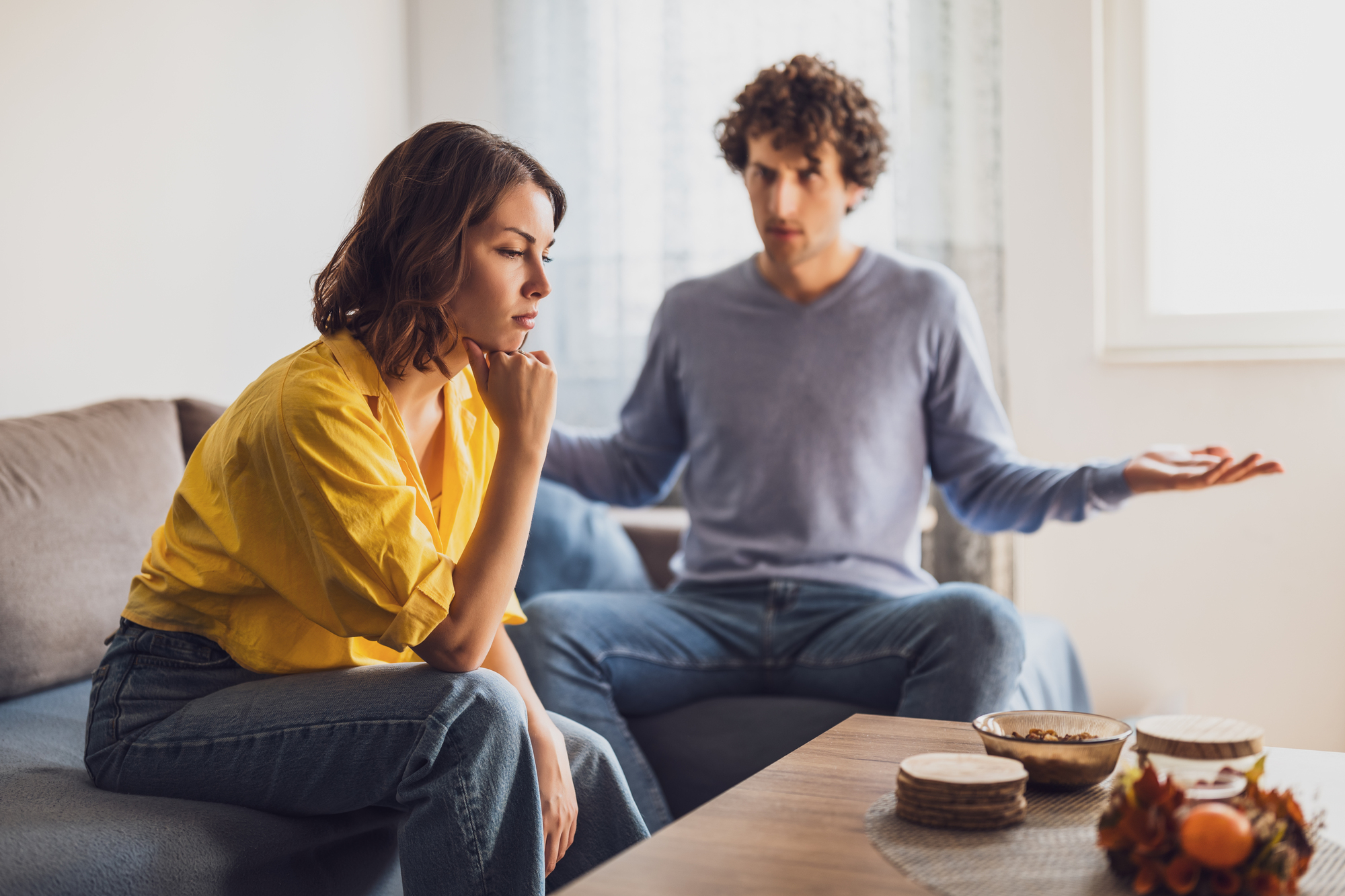 A woman in a yellow shirt sits thoughtfully on a couch, resting her chin on her hand. A man in a blue sweater sits beside her, gesturing with open arms. They appear to be having a serious conversation in a bright living room.