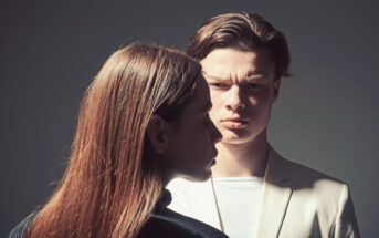 A young man and woman stand close together against a dark background. The man, wearing a white blazer, looks intensely at the woman, who has long brown hair and wears a dark jacket. The lighting highlights their facial expressions.