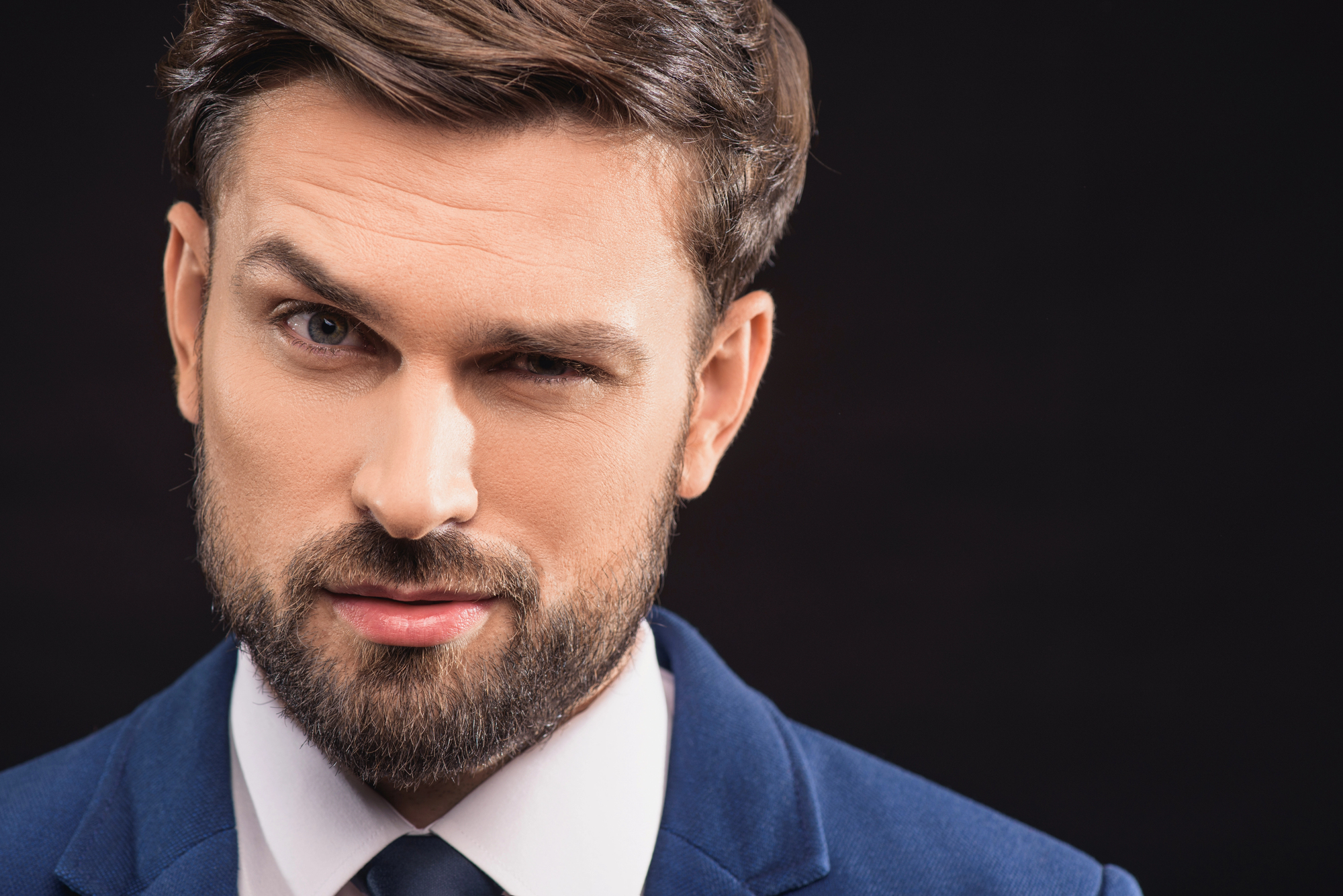 A man with a beard and mustache wearing a blue suit and white shirt is winking at the camera. He has short brown hair and is set against a dark background.