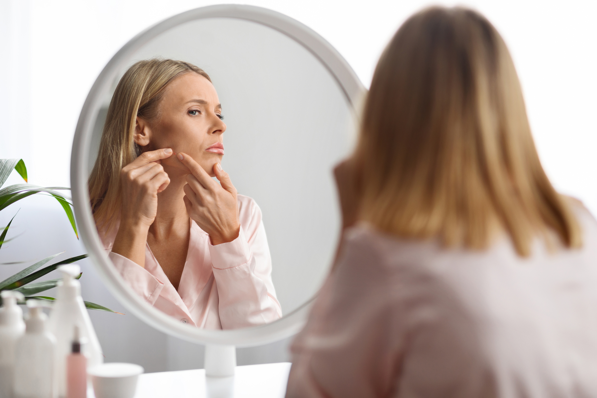 A woman in a pink blouse is examining her face in a round mirror. She is touching her cheek and looking thoughtful. There are skincare products on the table beside her. The room is bright, with a small plant visible in the background.