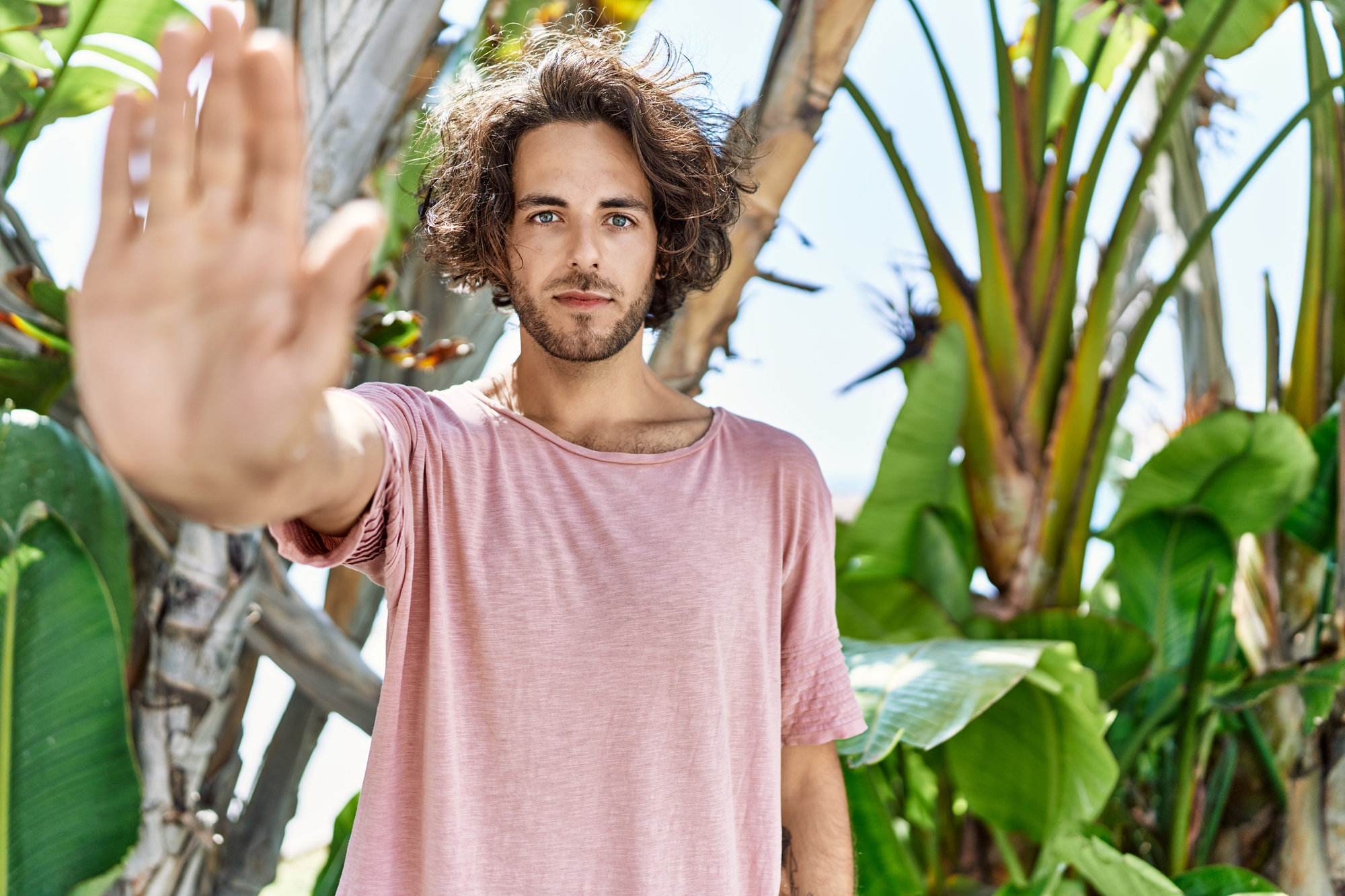 A person with curly hair wearing a pink shirt holds up their hand in a "stop" gesture. They stand outdoors in front of large green leaves and tropical plants, with a blue sky visible in the background.