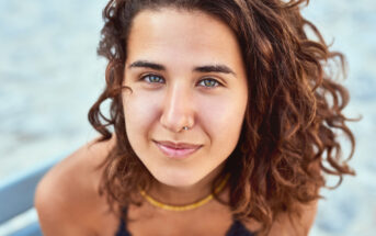 A person with curly brown hair and a gentle smile looks into the camera. They wear a nose ring and a necklace. The background is a soft blur, highlighting their features.