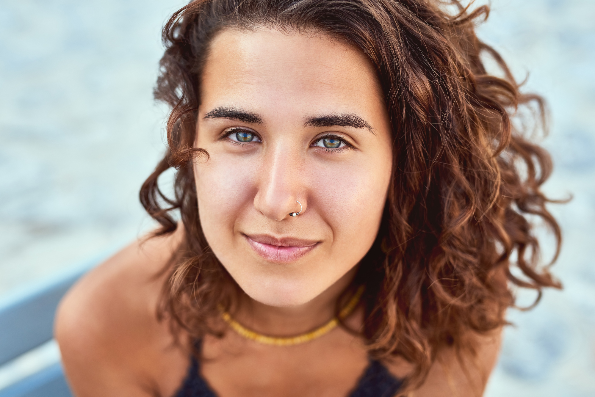 A person with curly brown hair and a gentle smile looks into the camera. They wear a nose ring and a necklace. The background is a soft blur, highlighting their features.