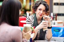 Two people sit at a table in a café, each holding a drink with whipped cream. The woman on the right is smiling, wearing a striped shirt and scarf, with curly hair. The person on the left is partially visible from the back.