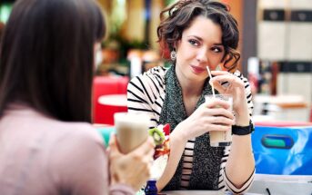 Two people sit at a table in a café, each holding a drink with whipped cream. The woman on the right is smiling, wearing a striped shirt and scarf, with curly hair. The person on the left is partially visible from the back.