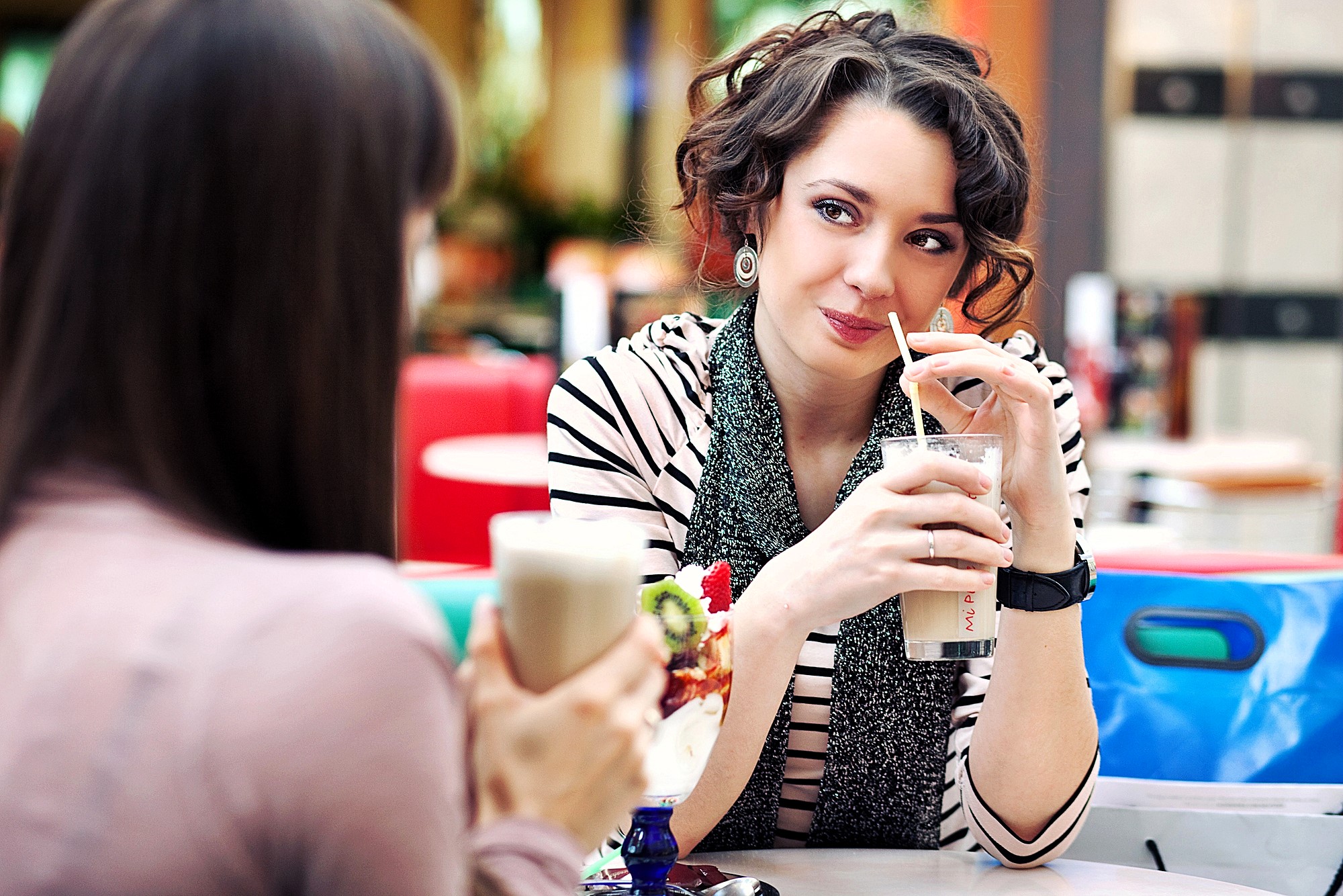 Two people sit at a table in a café, each holding a drink with whipped cream. The woman on the right is smiling, wearing a striped shirt and scarf, with curly hair. The person on the left is partially visible from the back.