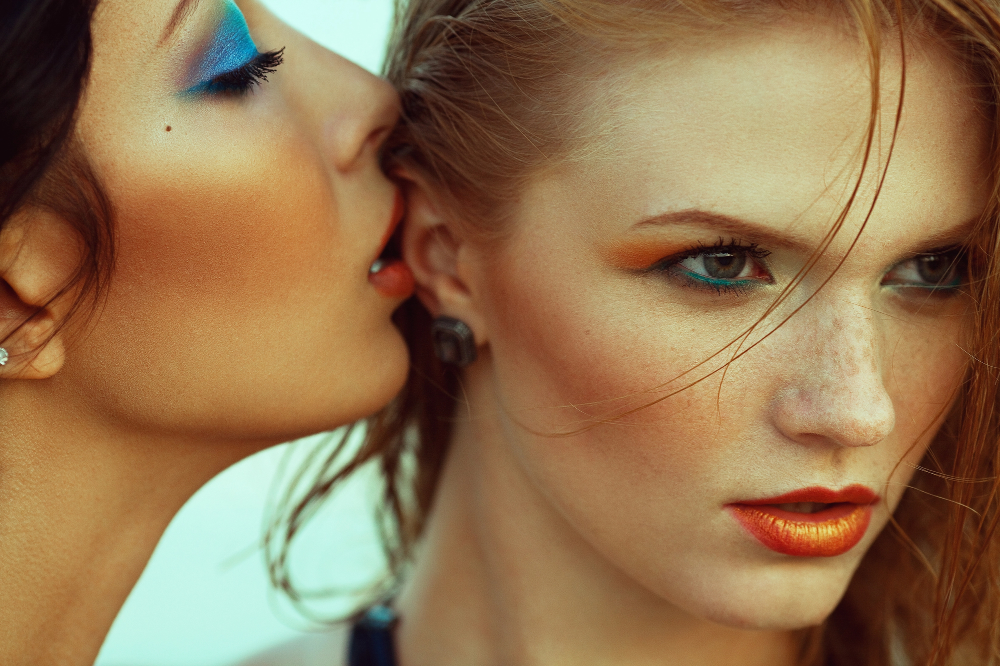 Two women with striking makeup are depicted closely together. One woman whispers into the other's ear, creating an intimate and enigmatic atmosphere. Their makeup features bold colors like blue and orange, highlighting their eyes and lips.