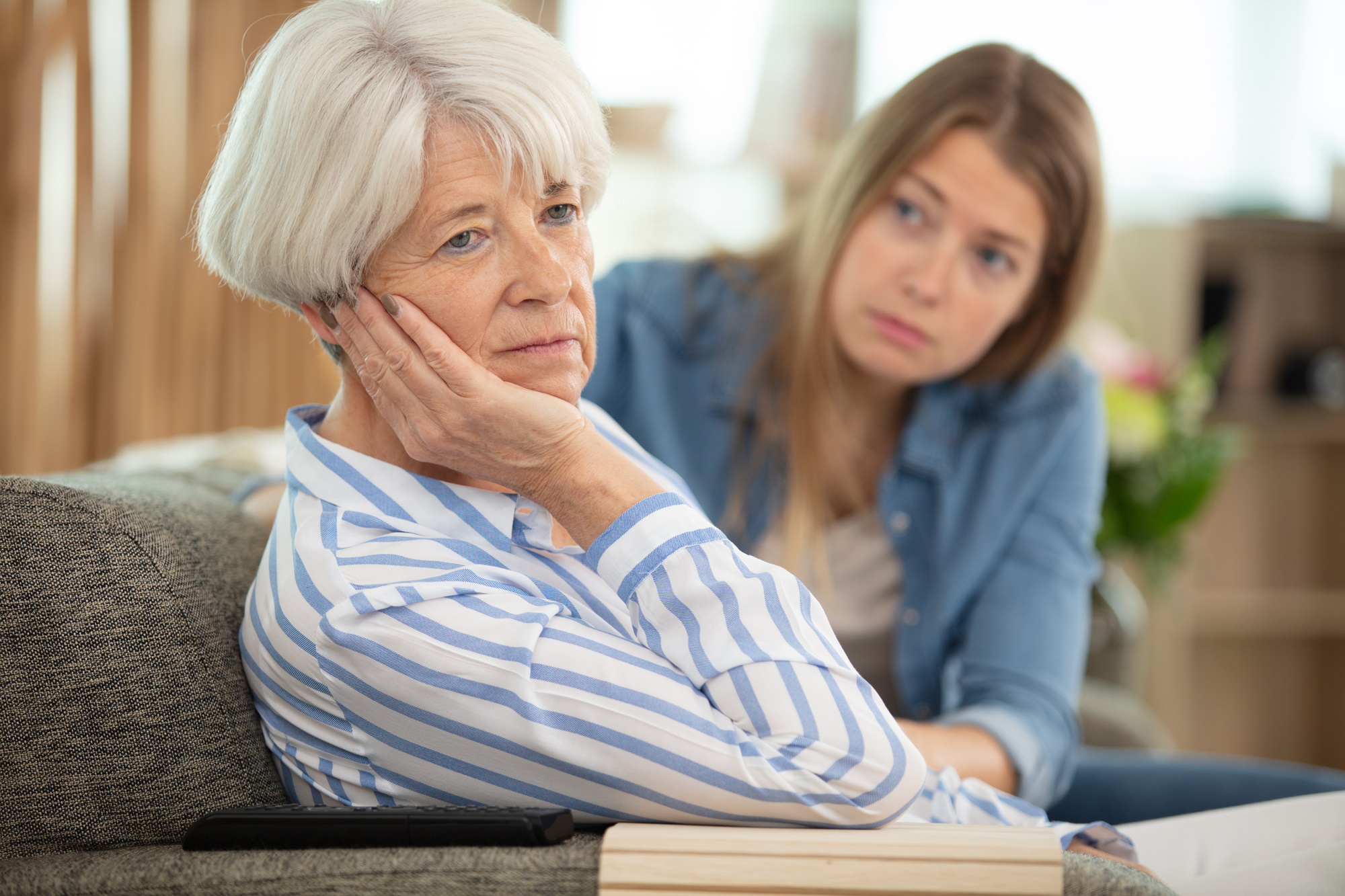 An older woman with short gray hair, wearing a striped shirt, looks thoughtful while sitting on a sofa. A younger woman with long hair sits beside her, appearing concerned. A book and a smartphone are on the sofa armrest.