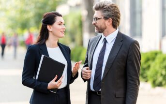 A man and a woman in business attire are walking outside and having a conversation. The woman is holding a folder, and they appear engaged in discussion. The background is blurred, suggesting a city or park setting.