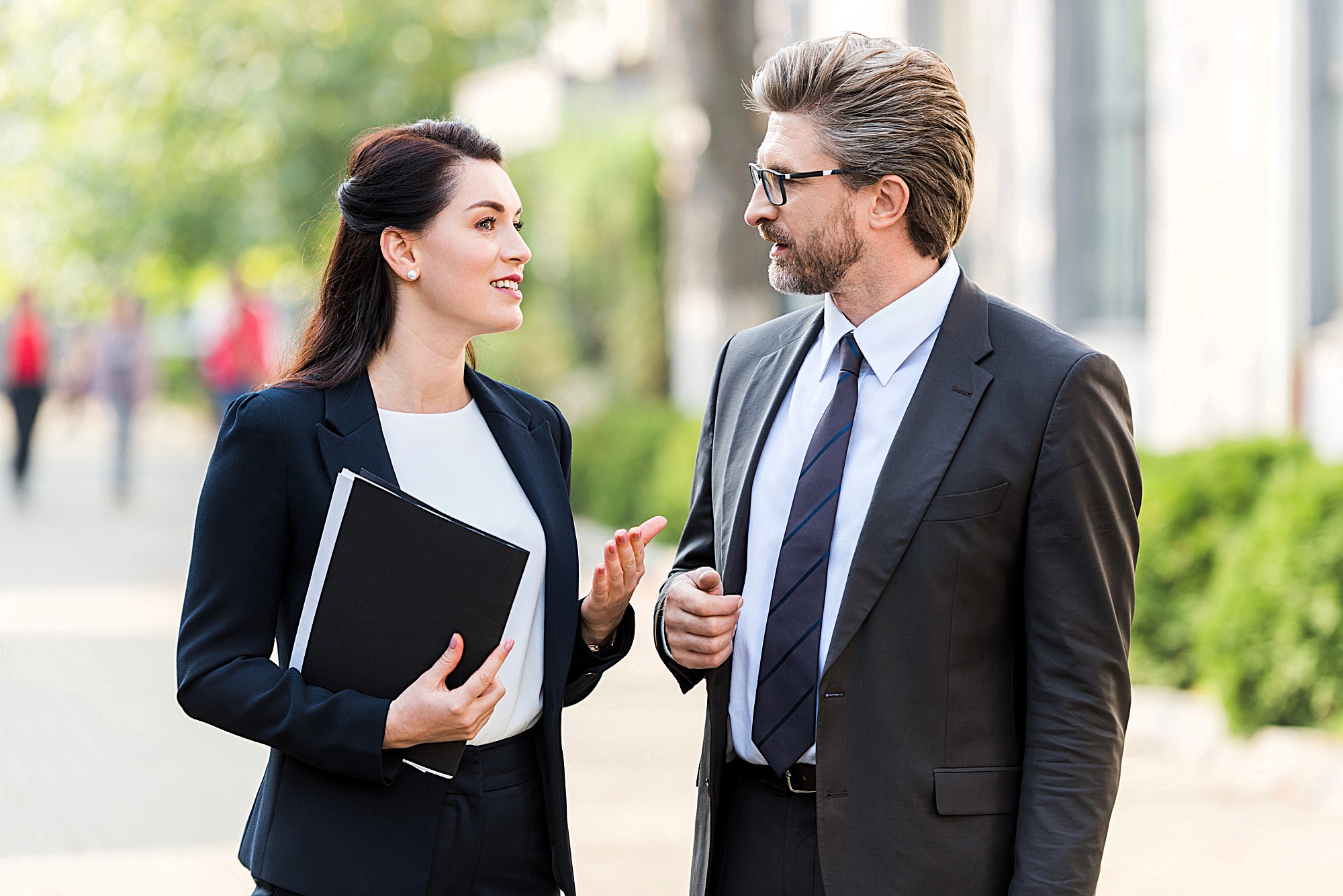 A man and a woman in business attire are walking outside and having a conversation. The woman is holding a folder, and they appear engaged in discussion. The background is blurred, suggesting a city or park setting.