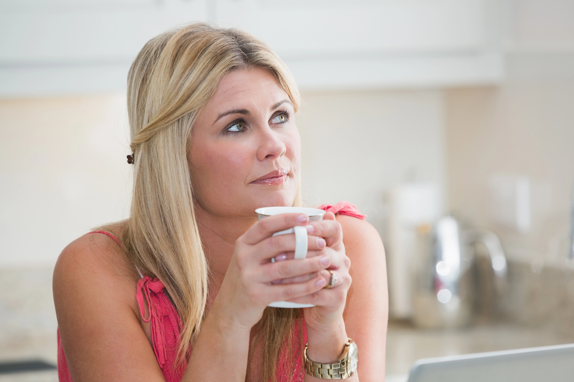 A woman with blonde hair sits in a kitchen, holding a white mug and gazing thoughtfully to the side. She is wearing a pink top and a gold watch. The background features a blurred kitchen countertop and appliances.